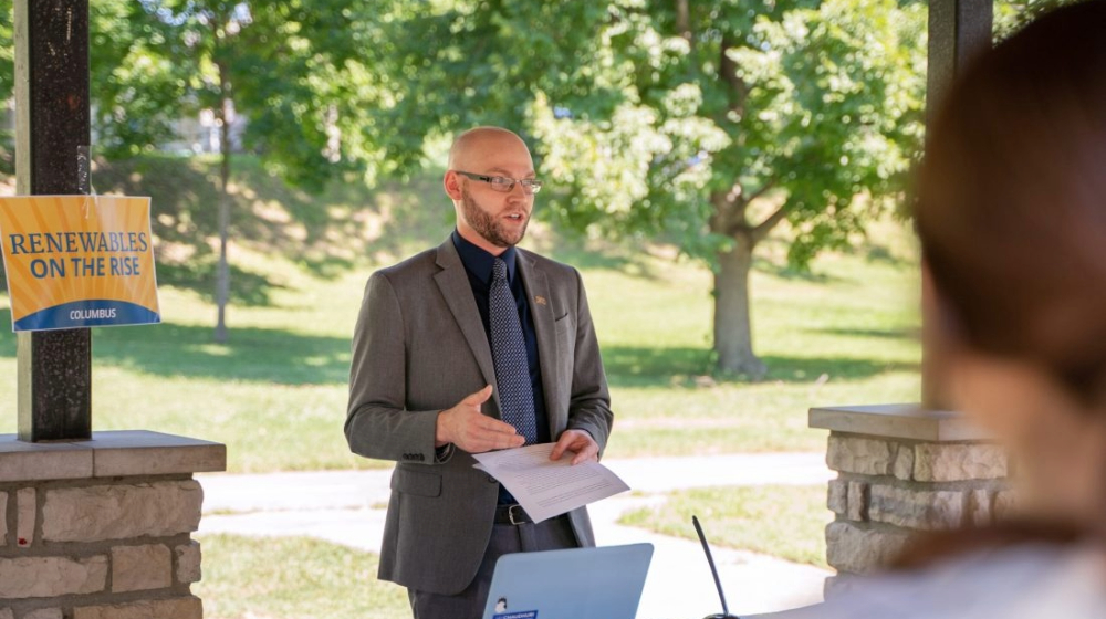 Gilbert Michaud pictured speaking at an event with trees and greenery in the background.